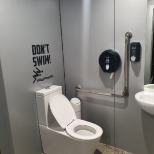 A modern bathroom with silver wet wall panels lining the wall, a white toilet, and a 'Don't Swim!' sign above the toilet. A grab bar, toilet paper dispenser, and soap dispenser are mounted on the wall.