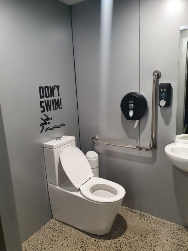 A modern bathroom with silver wet wall panels lining the wall, a white toilet, and a 'Don't Swim!' sign above the toilet. A grab bar, toilet paper dispenser, and soap dispenser are mounted on the wall.