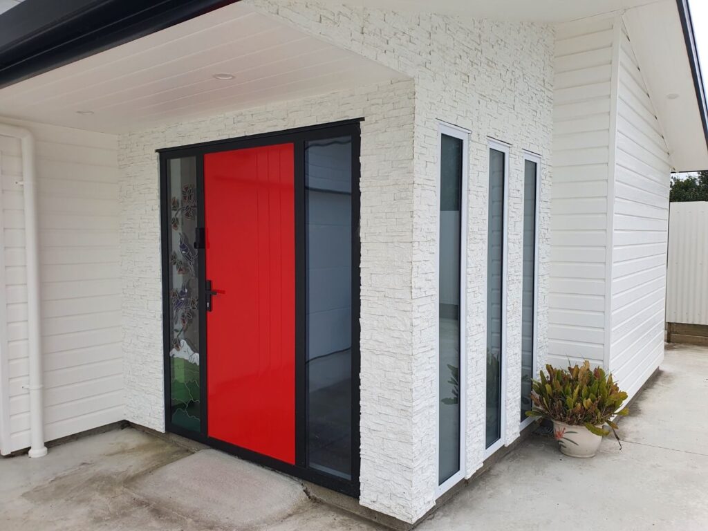 A modern house entrance with a red front door framed in black, set against a faux white stacked stone overclad wall. Either side of the front entrance exterior features a horizontal white weatherboard and tall narrow windows.