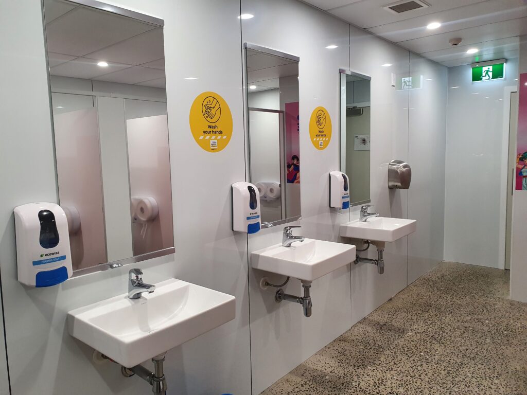 A commercial bathroom with white wet wall panels and negative detail. The room has three sinks, each with a mirror and a soap dispenser. Yellow signs between the mirrors remind users to wash their hands