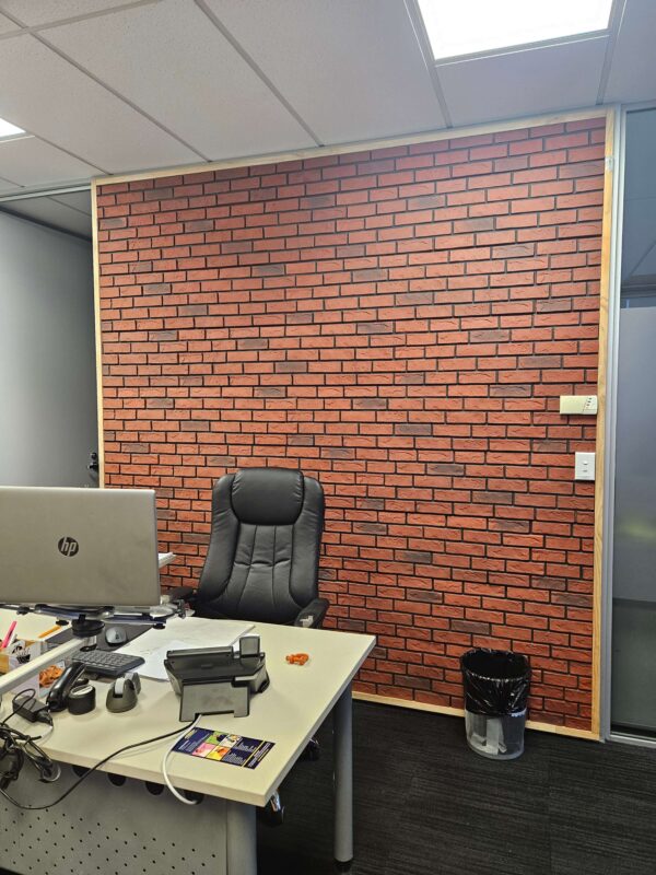 An office space with a faux red brick textured feature wall. In front of the wall is a desk with a laptop, office supplies, and a black swivel chair. A trash bin is placed to the right of the desk. The ceiling has white acoustic tiles and fluorescent lighting.