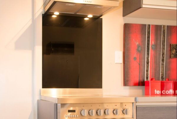 A modern kitchen with a stainless steel stove, black wet wall splash back, and a range hood above with lights. A red & black art canvas and containers are on the right above the bench.