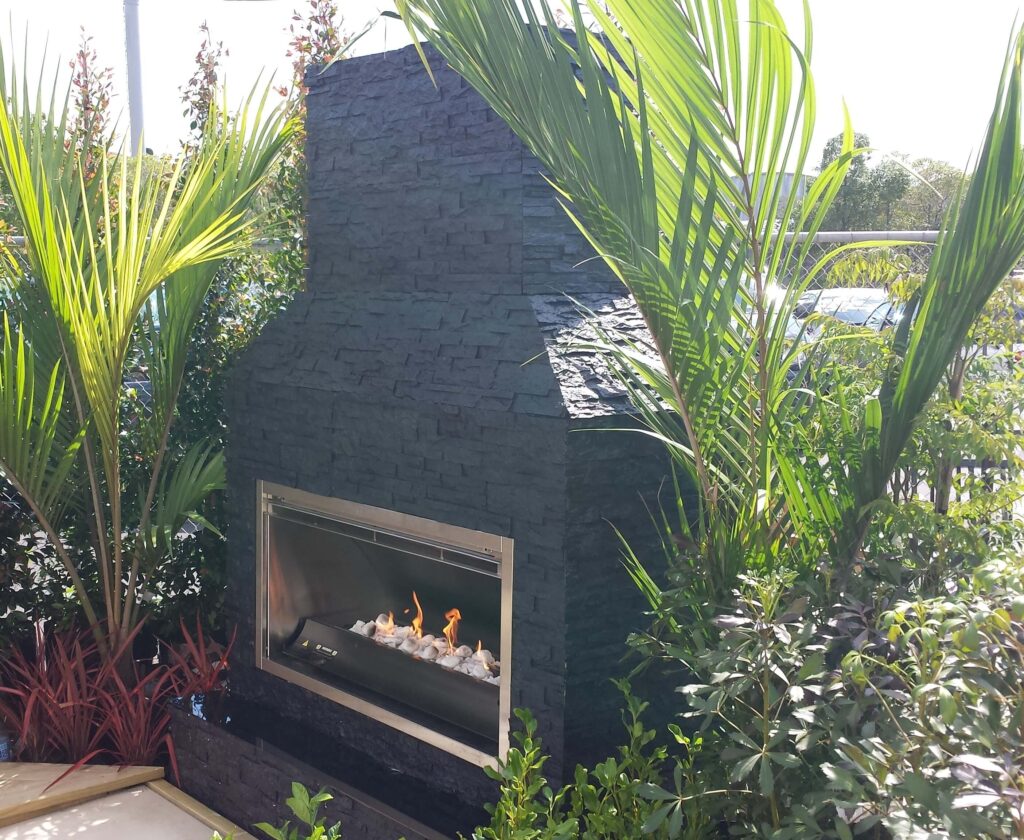 An outdoor fireplace with a faux black stacked stone finish, surrounded by lush green plants and palm fronds. The fireplace features a stainless steel firebox with flames visible over white decorative stones.