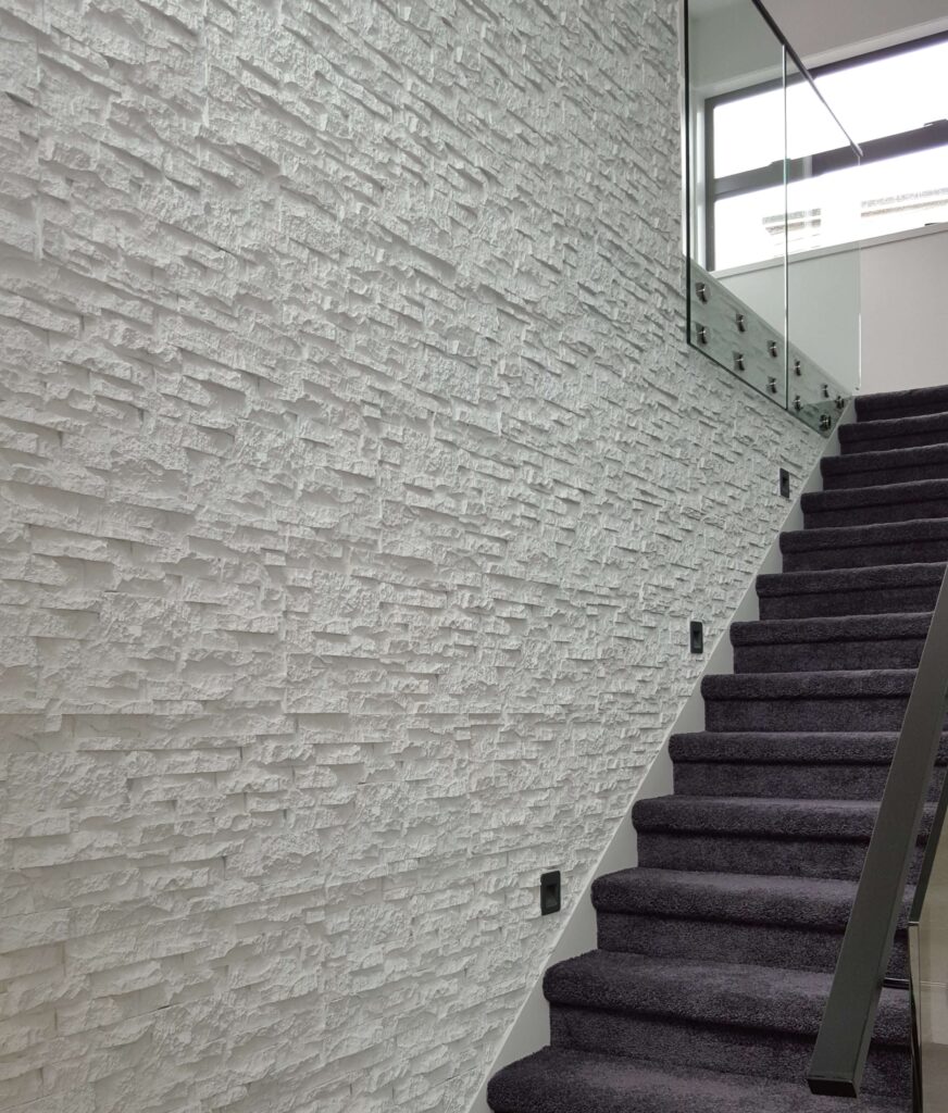 A staircase with dark grey carpeted steps and a modern glass railing. The adjacent wall features a faux white stack stone finish. Recessed black wall lights are evenly spaced along the faux stone wall.