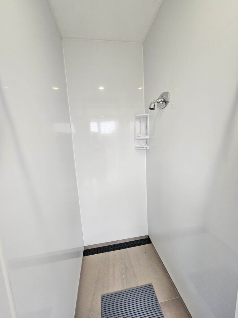 A shower with white wet wall panels, a small showerhead, and a light beige tiled floor with a black drain