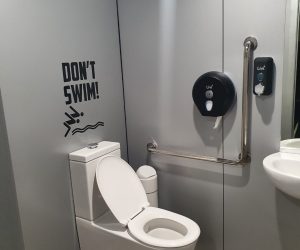 A modern bathroom with silver wet wall panels lining the wall, a white toilet, and a 'Don't Swim!' sign above the toilet. A grab bar, toilet paper dispenser, and soap dispenser are mounted on the wall.