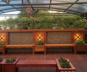 Outdoor seating area with a faux beige ledge stone feature wall, wooden benches with integrated planters, and warm lighting under a transparent pergola