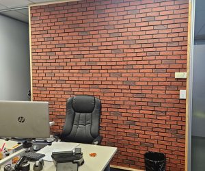 An office space with a faux red brick textured feature wall. In front of the wall is a desk with a laptop, office supplies, and a black swivel chair. A trash bin is placed to the right of the desk. The ceiling has white acoustic tiles and fluorescent lighting.