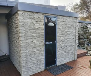 An outdoor structure with faux beige ledge stone over clad, featuring a black door with a sign that reads 'Steam Room, Keep Door Closed.' The building is set on a wooden deck, with natural stone wall in the surrounding area.