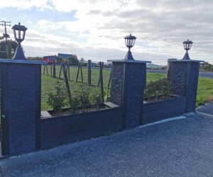 Outdoor planters filled with small shrubs that are in between three pillars. The pillars made out of Faux Stack Stone panels and are topped with lantern-style lights. The pillar and planter fence are positioned along a concrete driveway, with a grassy field and a fence in the background.