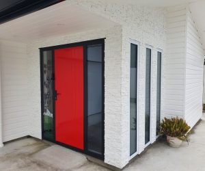 A modern house entrance with a red front door framed in black, set against a faux white stacked stone overclad wall. Either side of the front entrance exterior features a horizontal white weatherboard and tall narrow windows.