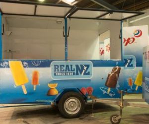 A blue mobile food trailer with white interior wet wall lining featuring 'Real NZ Since 1936' branding with images of ice creams and ice blocks, parked indoors with its serving window open.